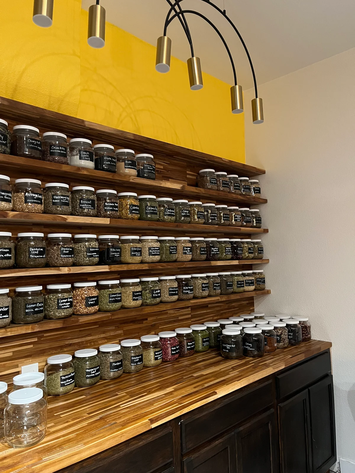 Rows of glass herb jars on wooden shelves against a yellow wall at Hearth and Seoul.